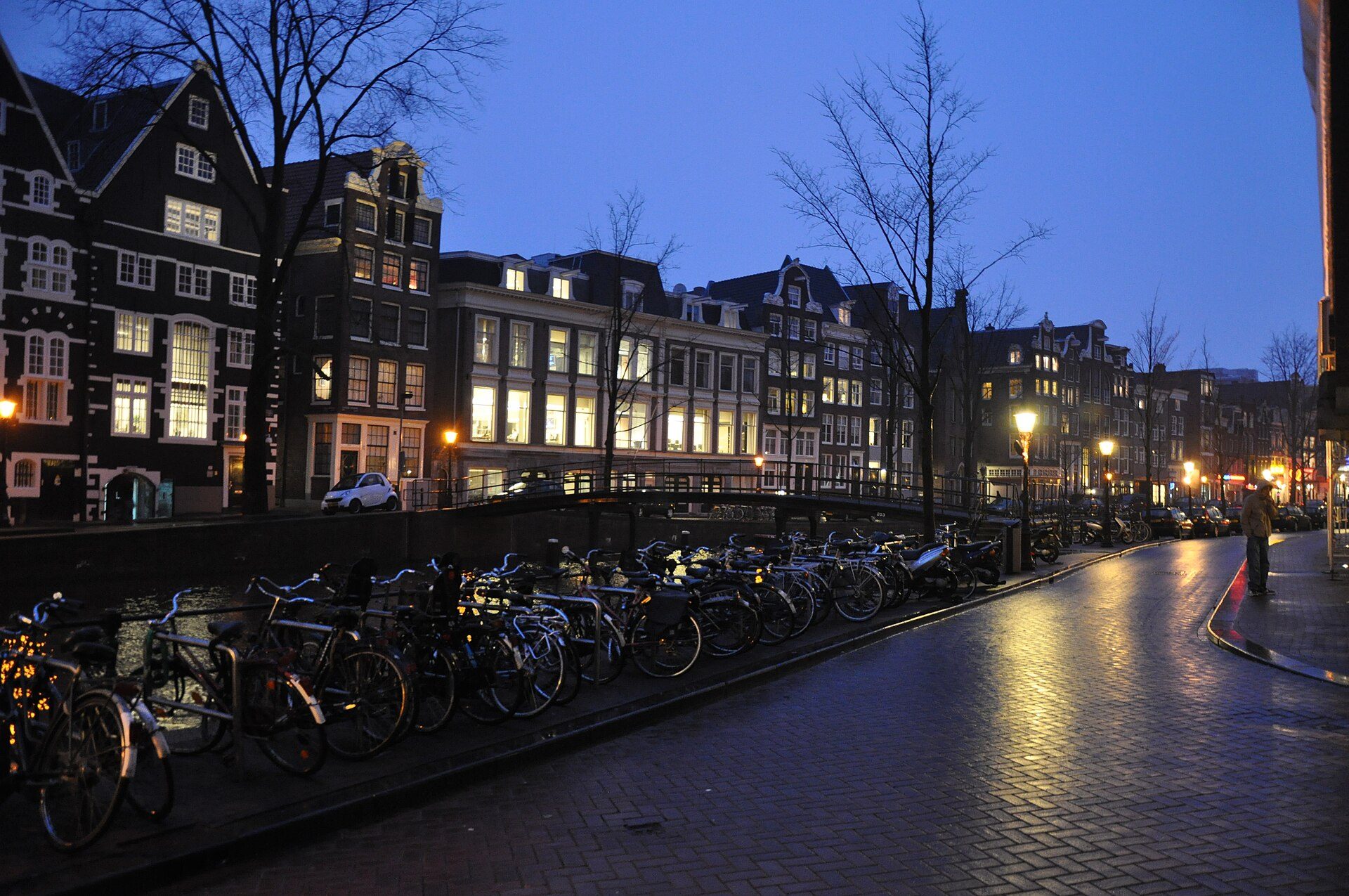 A small apartment block at blue hour, several windows lit, a person on the pavement below.